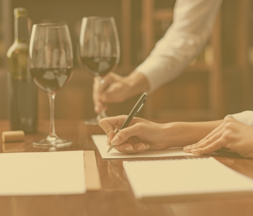 A wine critic writing tasting notes beside two glasses of red wine and an opened bottle