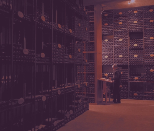 A worker at a lectern between tall racks of vintage-labelled bottles in a dark fine wine cellar